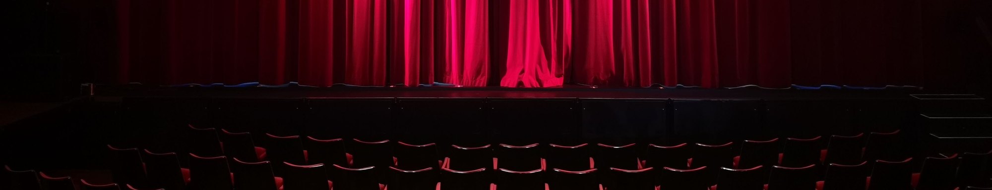 An empty theatre with lit red stage curtains above rows of seats